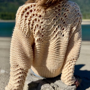 Może przedstawiać: Beżowy, ręcznie robiony sweter z unikalnym ażurowym wzorem na górnej części pleców i rękawach. Sweter ma prążkowaną fakturę na ciele i rękawach i jest wykonany z miękkiej, jasnej przędzy. Rękawy są długie, a sweter wydaje się być w średnim rozmiarze.