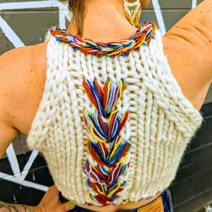 May include: Cream-coloured, hand-knitted halter top with a colourful braided neckline and back detail. The back features a vertical design with red, blue, yellow, and green threads. The top is paired with blue jeans.