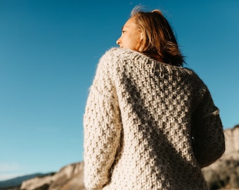 May include: A woman wearing a chunky knit cream sweater, standing with her back to the camera, looking off to the side. The background is a blue sky and a hillside.