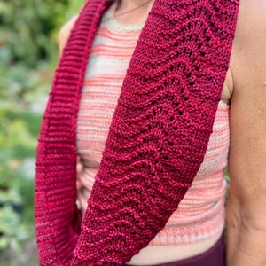 Puede incluir: Un bufanda infinita de punto en un tono rojo intenso. La bufanda presenta un diseño texturizado y calado en una parte de su longitud, con un patrón de punto acanalado en la otra. La bufanda está envuelta alrededor del cuello, mostrando su estilo y color.