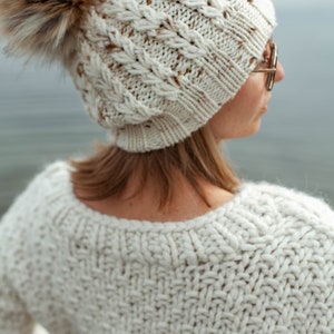 Puede incluir: Un gorro de punto blanco con un pomp&oacute;n de piel sint&eacute;tica marr&oacute;n. El gorro tiene un patr&oacute;n de punto de cable y lo lleva una persona con el pelo rubio.
