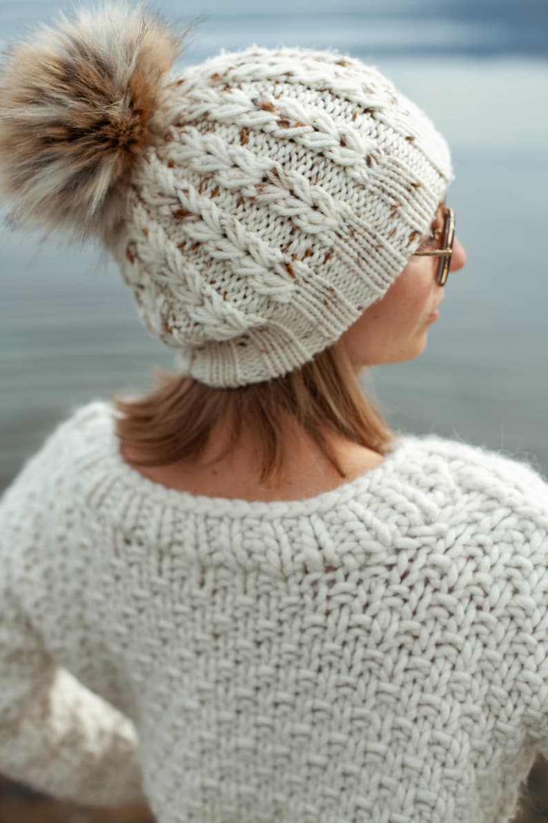 Puede incluir: Un gorro de punto blanco con un pomp&oacute;n de piel sint&eacute;tica marr&oacute;n. El gorro tiene un patr&oacute;n de punto de cable y lo lleva una persona con el pelo rubio.