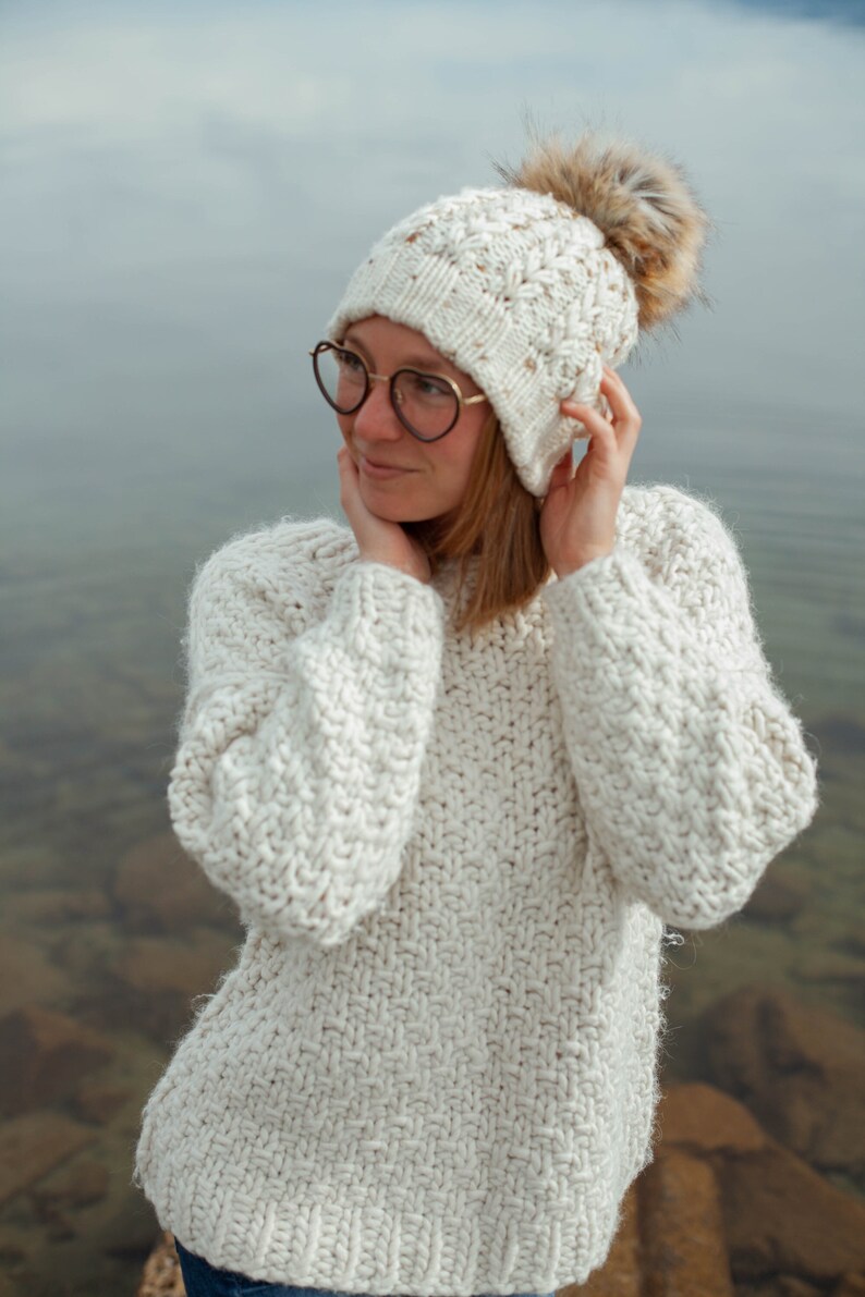 Puede incluir: Una mujer con un su&eacute;ter de punto blanco y un gorro de punto blanco con un pomp&oacute;n de piel marr&oacute;n. Lleva gafas redondas y tiene las manos en las mejillas.