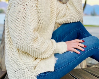 May include: A cream-colored, chunky knit sweater with a relaxed fit. The sweater has a round neckline and long sleeves. The model is wearing the sweater with blue jeans.