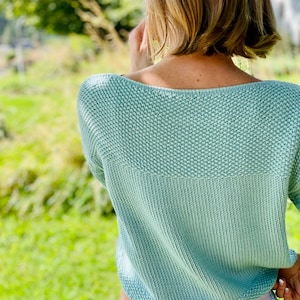 May include: A light green knitted jumper featuring a textured seed stitch pattern on the upper section and a smooth stockinette stitch on the lower body, with a wide neckline and short sleeves, worn over light-coloured bottoms with a leaf print.