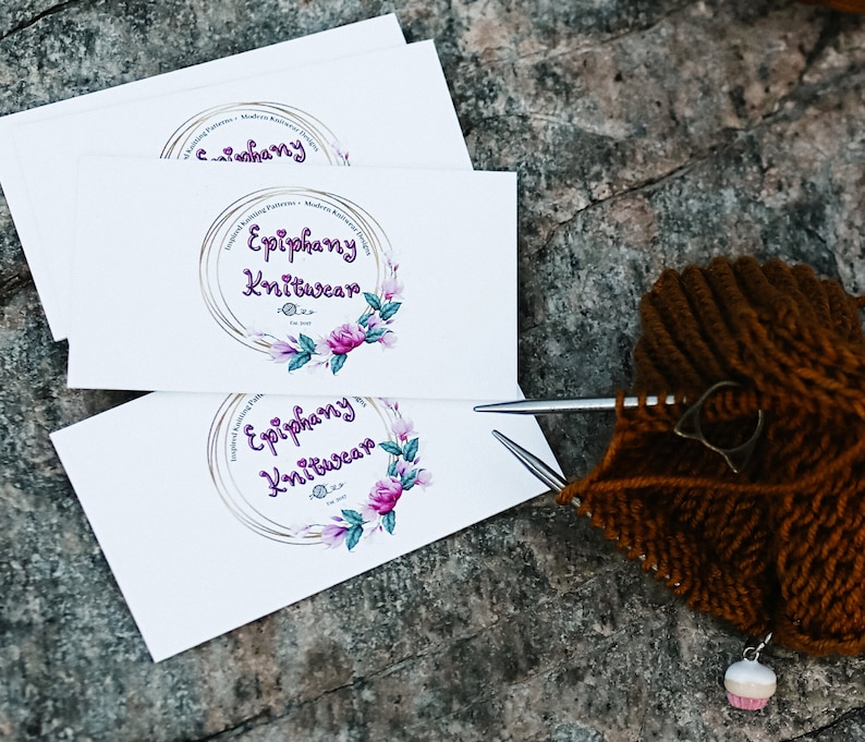 Może przedstawiać: Trzy białe wizyt&oacute;wki z tekstem "Epiphany Knitwear" i kwiatowym wzorem w odcieniach fioletu, r&oacute;żu i zieleni. Wizyt&oacute;wki są ułożone jedna na drugiej. W tle znajduje się brązowa dzianina.