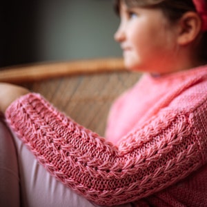 May include: A close-up of a pink knitted sweater with a cable knit pattern on the sleeves. The sweater is worn by a person sitting in a wicker chair.