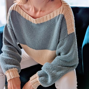 May include: A knitted sweater with a V-neckline. The sweater features a color-block design with a light blue body, a beige band across the chest, and beige accents on the neckline and cuffs. The sleeves are long and the fit is relaxed.