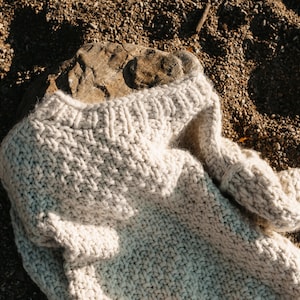 May include: A chunky knit cream-colored sweater lying on a bed of brown gravel.