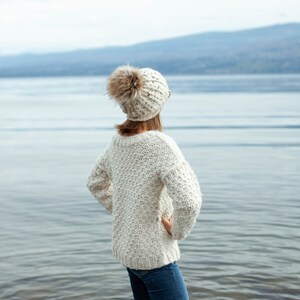 Puede incluir: Una mujer con un su&eacute;ter de punto blanco y una boina blanca con pomp&oacute;n, de pie junto a un lago y mirando hacia el agua.