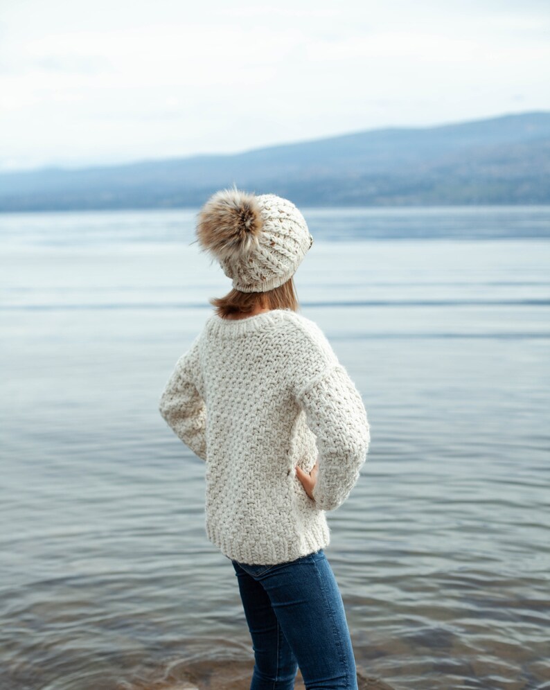 Puede incluir: Una mujer con un su&eacute;ter de punto blanco y una boina blanca con pomp&oacute;n, de pie junto a un lago y mirando hacia el agua.