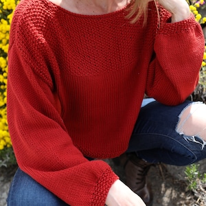May include: A red knitted sweater with a loose fit and long sleeves. The sweater has a simple, textured design.