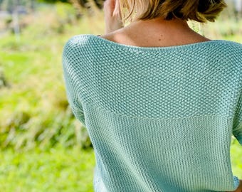 May include: A light mint green knit sweater featuring a textured seed stitch pattern across the upper back and shoulders, transitioning into a smooth stockinette stitch on the lower body, paired with light-colored patterned bottoms.