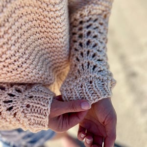 Könnte beinhalten: Nahaufnahme eines beigefarbenen Strickpullovers. Der Pullover hat ein strukturiertes Strickmuster am Körper und dekorative Lochmusterdetails an den Ärmeln. Die Ärmel sind lang und enden am Handgelenk. Der Pullover ist aus weichem, warmem Material.
