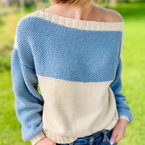 以下が含まれることがあります： 肩出しの広いネックラインを持つ、青とクリーム色のニットセーター。セーターは質感のあるニットで、ゆったりとしたフィット感です。