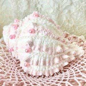 May include: A decorative seashell, white with pink accents and beaded embellishments, rests on a pink and white crocheted doily. A small seashell and a pink starfish are also present, with a floral arrangement in the background.