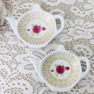 May include: Two white ceramic tea bag holders shaped like teapots. The holders have a pink, green, and yellow floral design with a pink rose in the center.