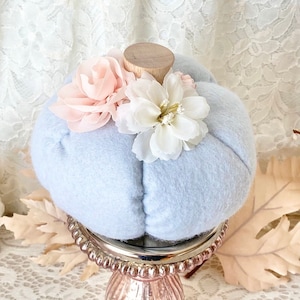 May include: A light blue felt pumpkin with white and pink fabric flowers on top. The pumpkin is sitting on a gold metal stand with a round top and three legs.