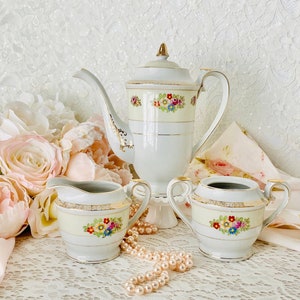 May include: A white porcelain tea set with a floral design. The set includes a teapot, creamer, and sugar bowl. The teapot has a gold trim and a floral design with pink, yellow, and blue flowers. The creamer and sugar bowl have a similar design. A string of pearls is in front of the tea set.