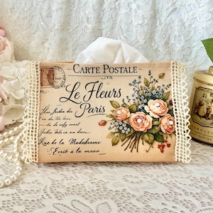 May include: A rectangular fabric tissue box cover featuring a vintage postcard design with the text "CARTE POSTALE" and "Le Fleurs Paris." The cover is decorated with a floral bouquet illustration, cursive script, and cream-colored lace trim on the sides.