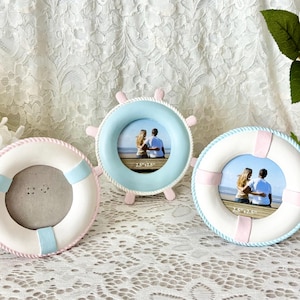 May include: Three decorative picture frames in the shape of a life preserver and a ship's wheel. The frames are white with blue and pink accents. The frames are empty and ready for photos.