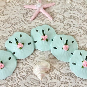 May include: A collection of sea-themed decorations, including five light blue sand dollars with pink floral accents, a pink starfish, and a white seashell. The items are arranged on a white lace surface, creating a coastal aesthetic.