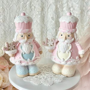 May include: Two decorative figurines of a nutcracker dressed in pink and white, with a cupcake-shaped hat. The nutcrackers have white beards, blue pants, and are holding a small tray. They are standing on a white pedestal with a lace doily.