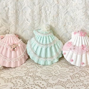 May include: Three decorative seashell-shaped trinket boxes in pastel colors. The boxes are adorned with lace trim, pearls, and floral accents. The shells are pink, mint green, and white, and are arranged on a lace tablecloth.
