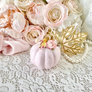 May include: A pink glitter pumpkin with a small pink flower on top, surrounded by a bouquet of pink roses and peonies, a gold glitter leaf, and a pearl necklace on a white lace background.