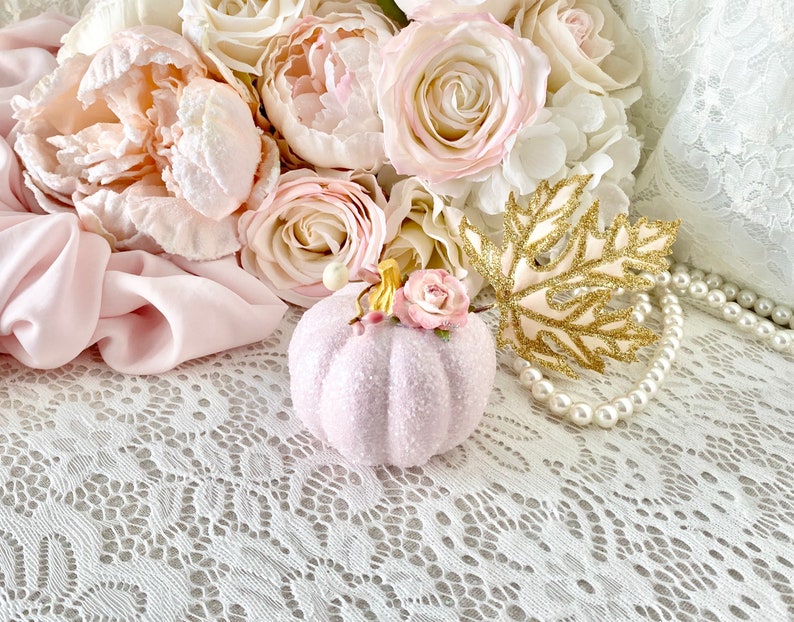 May include: A pink glitter pumpkin with a small pink flower on top, surrounded by a bouquet of pink roses and peonies, a gold glitter leaf, and a pearl necklace on a white lace background.