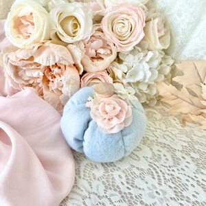 May include: A light blue felt pumpkin with a pink felt flower on top. The pumpkin is sitting on a white lace tablecloth with a bouquet of pink and white flowers in the background.