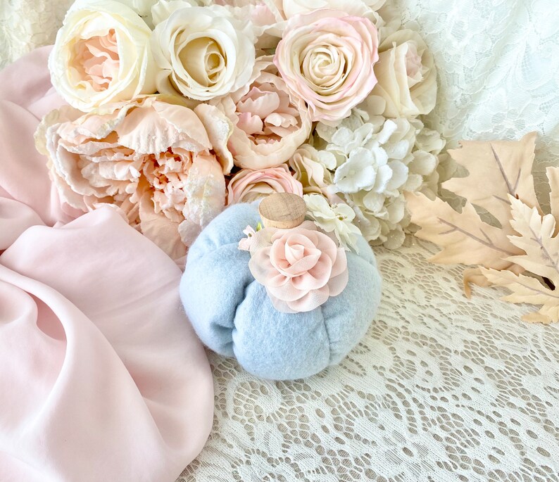 May include: A light blue felt pumpkin with a pink felt flower on top. The pumpkin is sitting on a white lace tablecloth with a bouquet of pink and white flowers in the background.