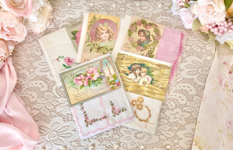 May include: Assortment of vintage handkerchiefs with floral and angel designs. The handkerchiefs are white with pink and blue embroidery. The handkerchiefs are displayed on a white lace tablecloth with pink roses.