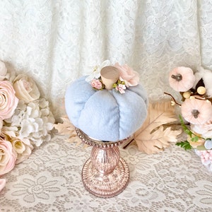 May include: A light blue velvet pumpkin with pink and white floral accents sits on a gold pedestal. The pumpkin is a fall decoration.