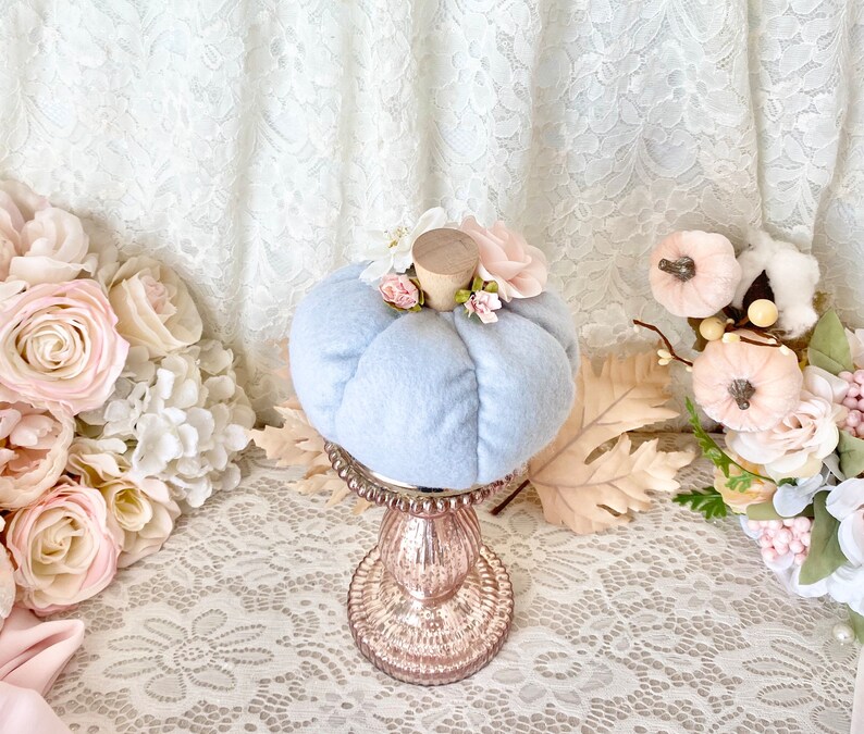 May include: A light blue velvet pumpkin with pink and white floral accents sits on a gold pedestal. The pumpkin is a fall decoration.