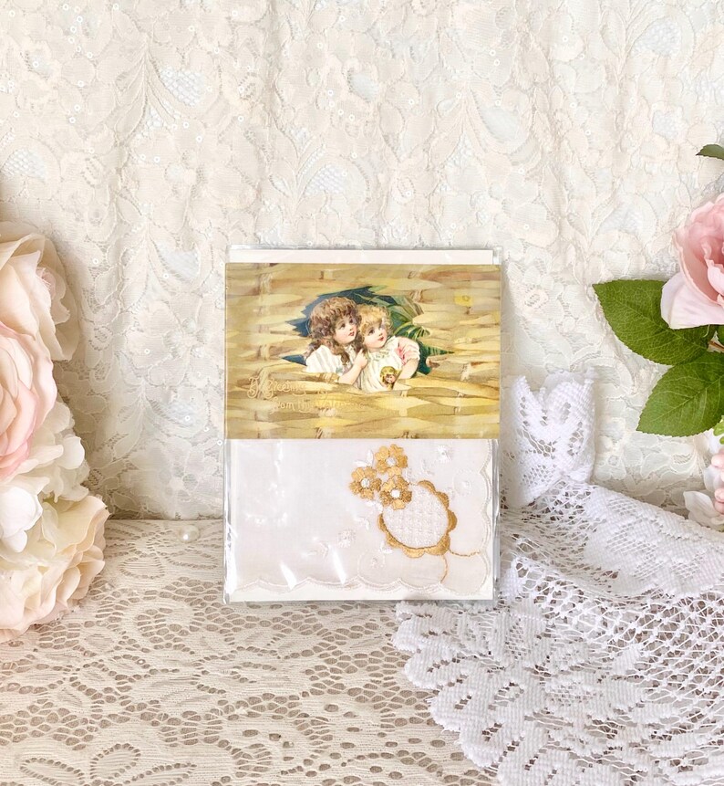 May include: A white handkerchief with gold embroidery of two flowers in a circle. The handkerchief is packaged in a clear plastic sleeve with a vintage postcard featuring two children in a field of wheat.