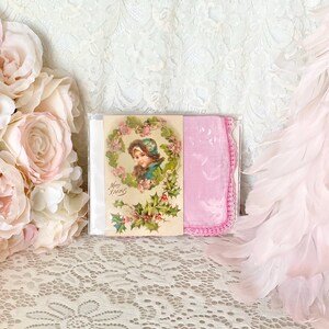 May include: A pink and white Christmas card with a vintage illustration of a girl in a wreath. The card is packaged with a pink handkerchief with a crocheted border.