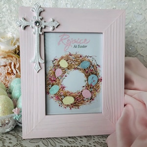 May include: A pink wooden picture frame with a decorative silver cross attached. The framed image features an Easter wreath with pastel-colored eggs and the words "Rejoice At Easter". The frame is set against a lace background.