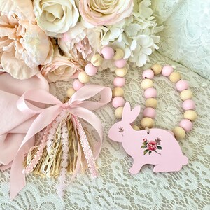 Pu&ograve; includere: Un ornamento a forma di coniglietto in legno rosa con un design floreale, abbinato a una ghirlanda di perline rosa e in legno naturale. &Egrave; presente anche un nappa rosa con dettagli in pizzo e nastro, insieme a un bouquet di fiori rosa e crema.