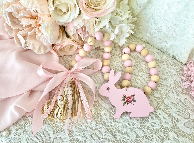 Pu&ograve; includere: Un ornamento a forma di coniglietto in legno rosa con un design floreale, abbinato a una ghirlanda di perline rosa e in legno naturale. &Egrave; presente anche un nappa rosa con dettagli in pizzo e nastro, insieme a un bouquet di fiori rosa e crema.