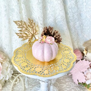 May include: A pink glitter pumpkin with a gold leaf, a pink rose, and a pine cone on top. The pumpkin is sitting on a gold doily on a white cake stand.