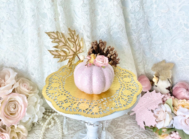 May include: A pink glitter pumpkin with a gold leaf, a pink rose, and a pine cone on top. The pumpkin is sitting on a gold doily on a white cake stand.