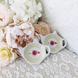 May include: Two white ceramic tea bag holders shaped like teapots with pink and yellow floral designs. The holders are sitting on a white lace surface.
