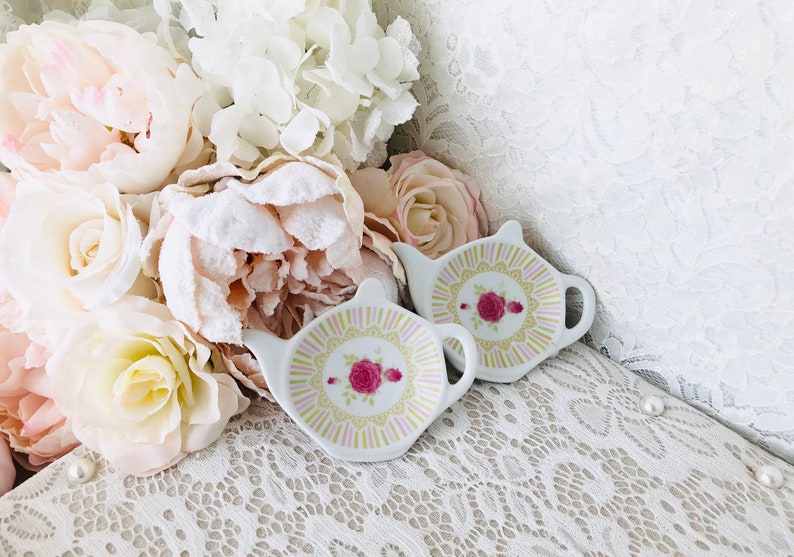 May include: Two white ceramic tea bag holders shaped like teapots with pink and yellow floral designs. The holders are sitting on a white lace surface.