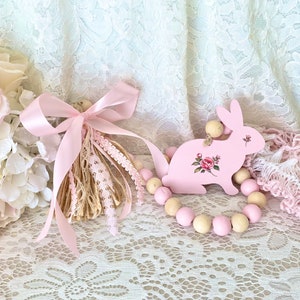 Pu&ograve; includere: Un coniglietto di legno rosa con un motivo floreale e una ghirlanda di perline di legno. Un nappa rosa con pizzo e nastro &egrave; a sinistra. Lo sfondo include tessuto di pizzo e fiori rosa.