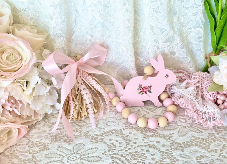 Pu&ograve; includere: Un coniglietto di legno rosa con un motivo floreale e una ghirlanda di perline di legno. Un nappa rosa con pizzo e nastro &egrave; a sinistra. Lo sfondo include tessuto di pizzo e fiori rosa.