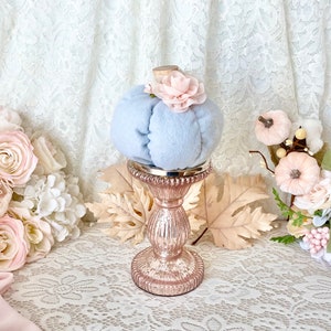 May include: A light blue velvet pumpkin with a pink flower on top, sitting on a rose gold pedestal. The pumpkin is surrounded by fall leaves and pink flowers.