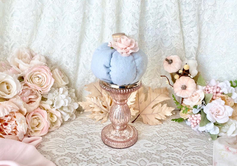 May include: A light blue velvet pumpkin with a pink flower on top, sitting on a rose gold pedestal. The pumpkin is surrounded by fall leaves and pink flowers.