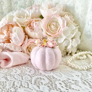 May include: A pink glittery pumpkin with a small pink flower on top, surrounded by a bouquet of white and pink flowers and a pearl necklace on a white lace background.