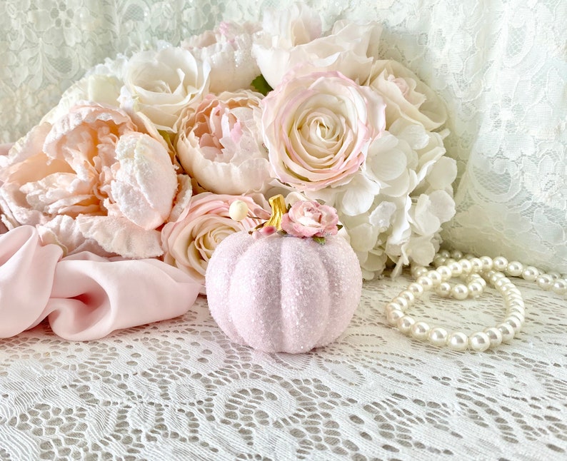 May include: A pink glittery pumpkin with a small pink flower on top, surrounded by a bouquet of white and pink flowers and a pearl necklace on a white lace background.